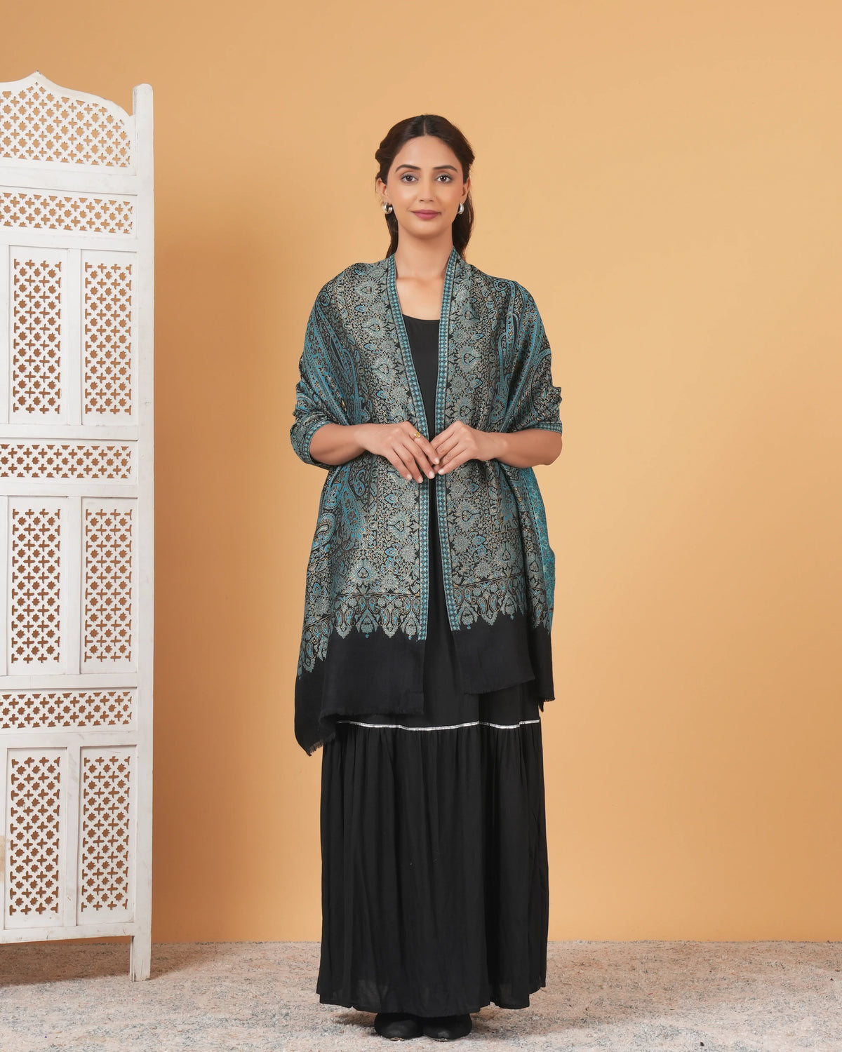 Woman wearing a black outfit with a green patterned shawl against a beige wall.