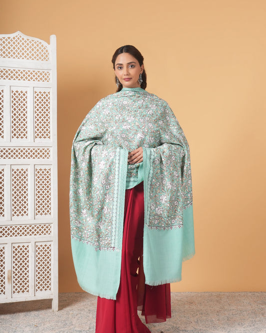 Woman wearing a light blue embroidered shawl over a red outfit against a beige wall.