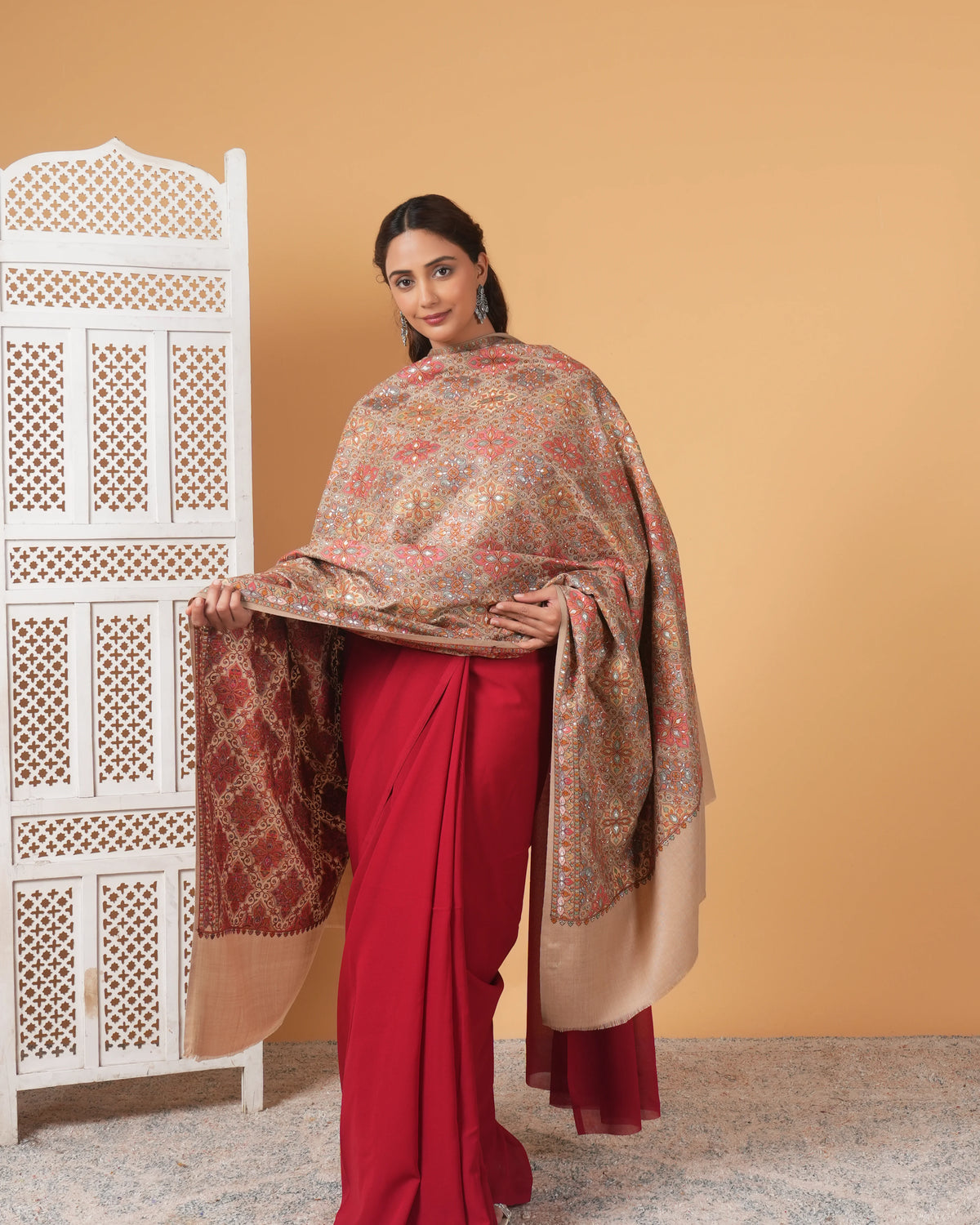 Woman wearing a red saree with a patterned shawl against a beige wall.