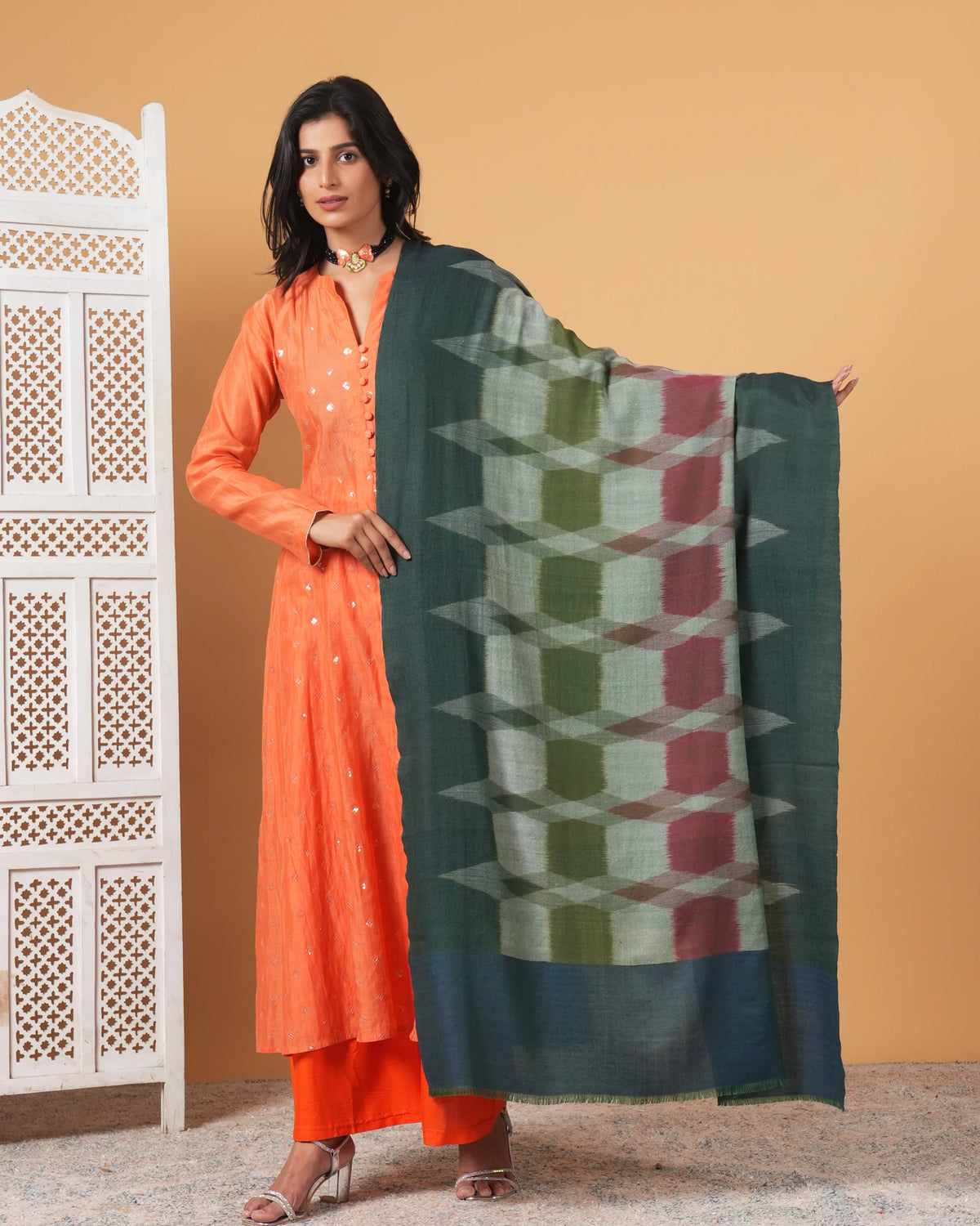 Woman in orange traditional outfit holding a patterned green scarf against a beige wall.