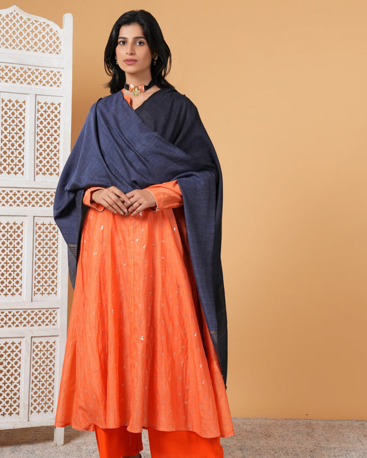 Woman wearing an orange outfit with a navy blue shawl against a beige wall.