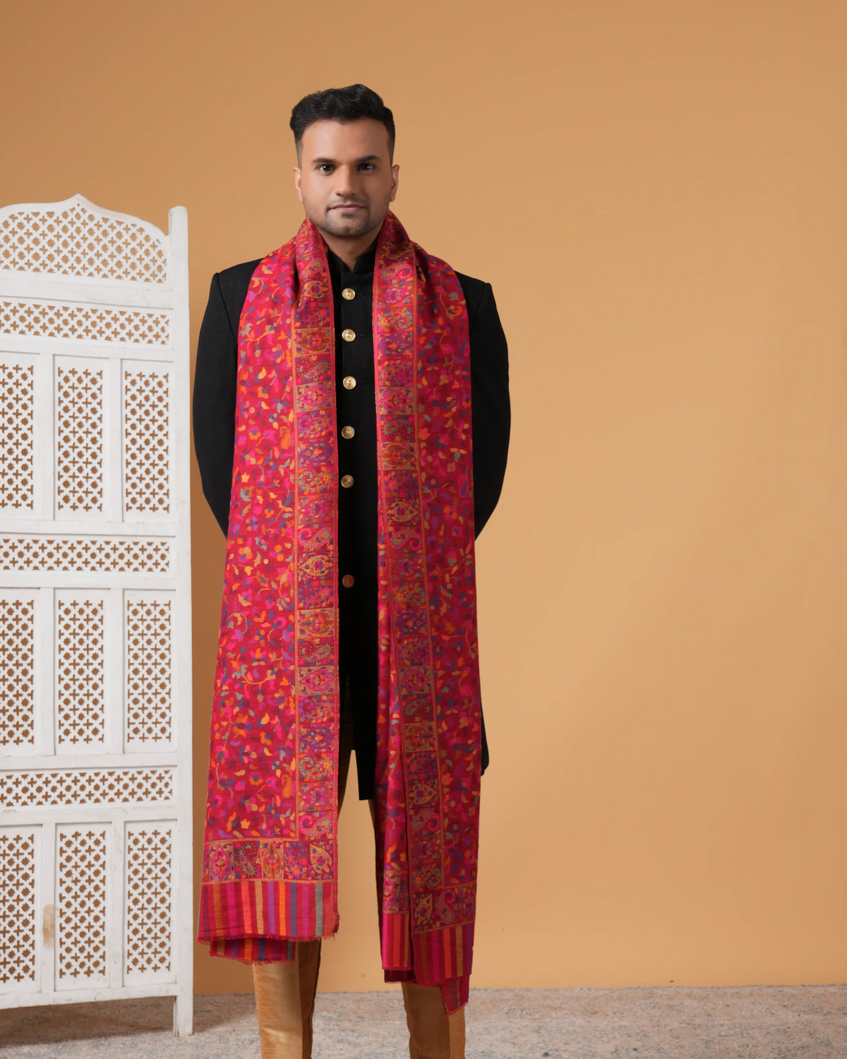 Man wearing a red patterned scarf with a black outfit against an orange wall.