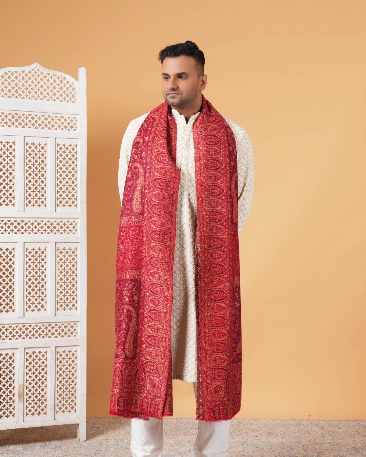 Man wearing a red patterned scarf and white outfit against a beige wall with a decorative white cabinet.