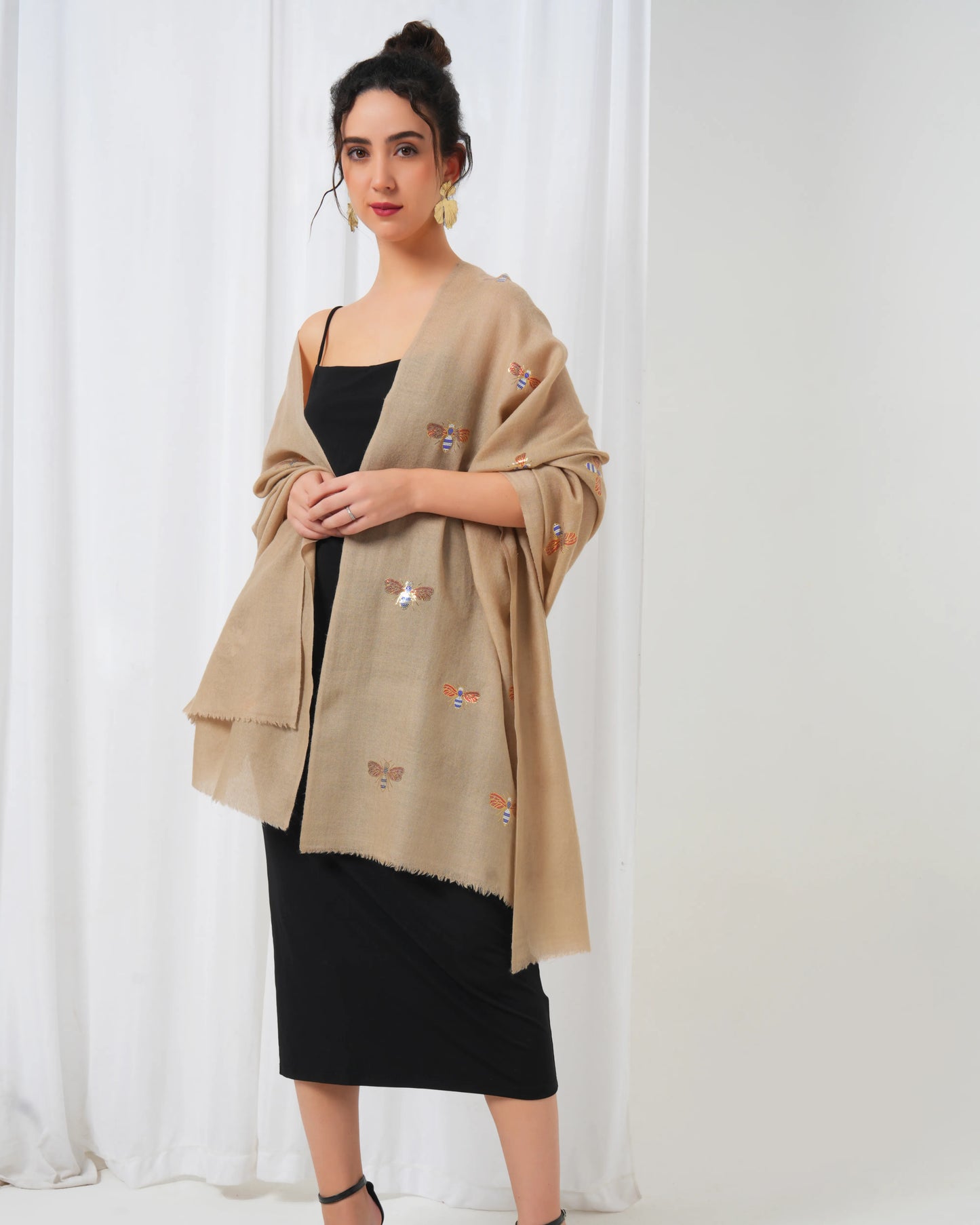 Woman wearing a beige shawl with embroidery over a black dress against a white curtain background