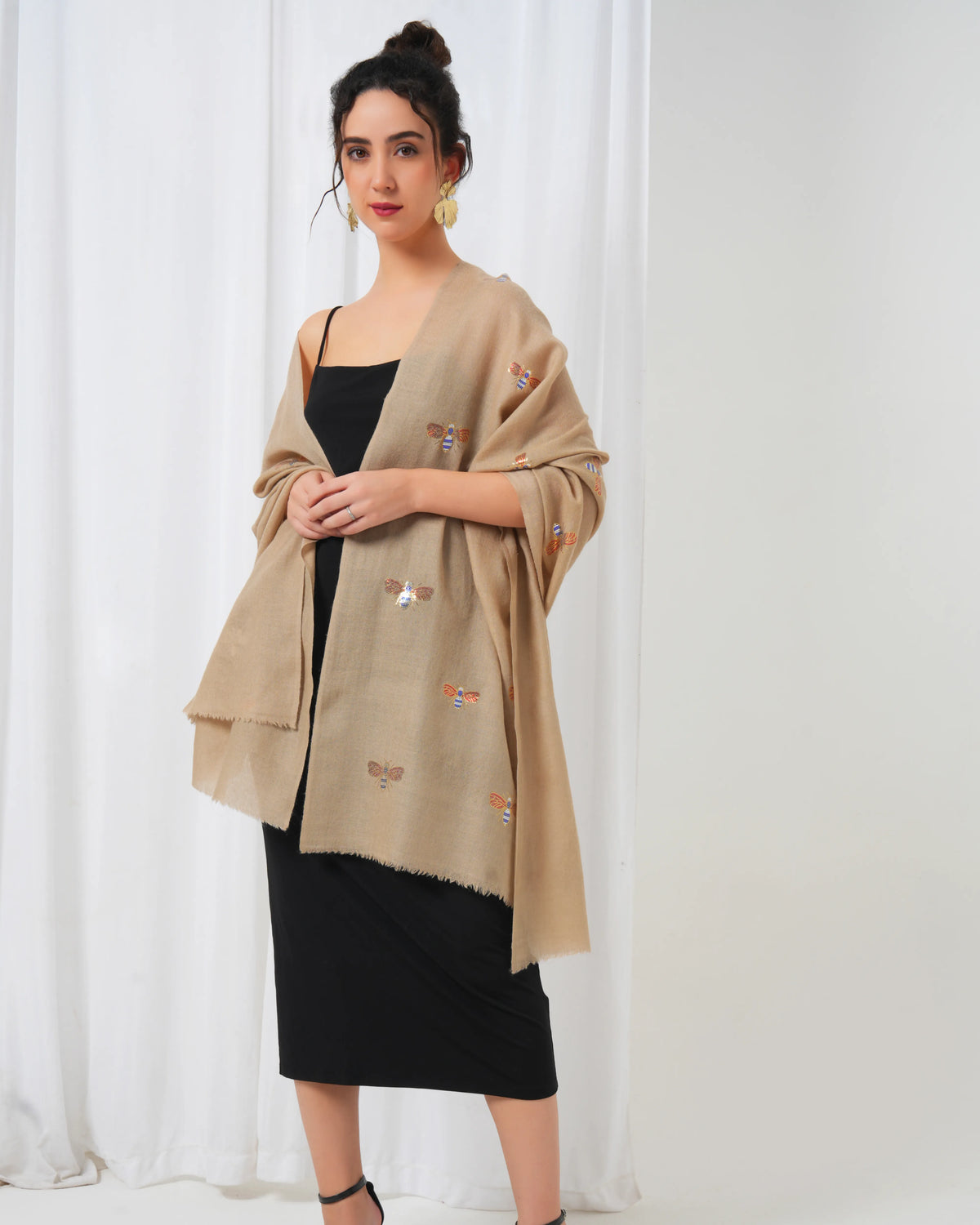 Woman wearing a beige shawl with embroidery over a black dress against a white curtain background